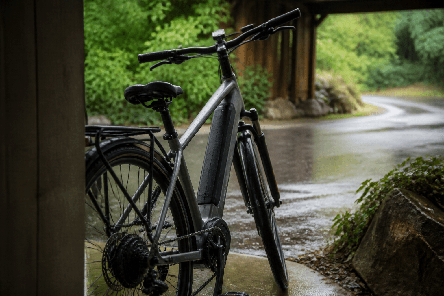 An e-bike parked under a shelter after riding in the rain, with water droplets on the frame and a wet road in the background.